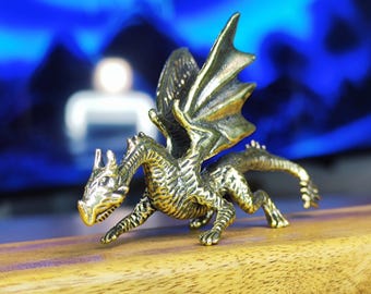 May include: A detailed brass dragon figurine with outstretched wings and textured scales. The dragon is posed on a wooden surface, with a blurred blue background. The figurine is a fantasy-themed collectible.