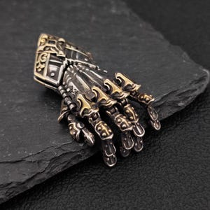 May include: A detailed, silver-toned metal hand pendant with gold-colored accents. The articulated fingers and intricate design suggest a medieval or fantasy theme. The pendant is displayed on a dark, textured surface.