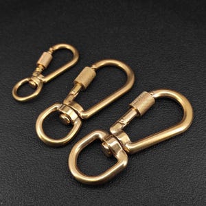 Könnte beinhalten: Drei goldfarbene, drehbare Karabinerhaken aus Metall in verschiedenen Größen. Jeder Karabiner verfügt über einen sicheren Schraubverschluss mit einer texturierten, gerändelten Hülse und eine glatte, abgerundete Basis zum Befestigen von Riemen.