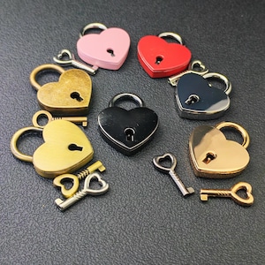 Puede incluir: Colección de candados en forma de corazón en varios colores, incluyendo rosa, rojo, dorado, negro y plateado. Cada candado tiene una llave a juego. Los candados están dispuestos en un patrón circular.