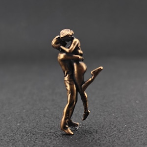 May include: A bronze-colored sculpture depicting a couple embracing in a passionate pose. The man is holding the woman, who is leaning back with one leg raised. The sculpture has a smooth finish and is set against a dark background.