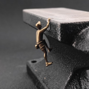 May include: A small bronze-colored figurine of a climber scaling the side of a heavy, black cast iron anvil. The climber is depicted in a dynamic pose, gripping the edge of the anvil's flat top surface with one hand while wearing climbing gear.