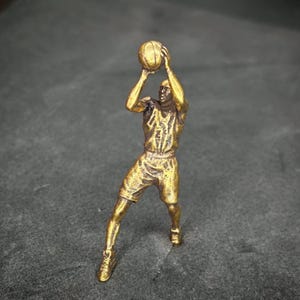 Può includere: Piccola statuetta dorata di un giocatore di basket in posizione di tiro. La figura indossa una maglia e pantaloncini, con la palla tenuta sopra la testa. I dettagli della figura sono visibili, comprese le scarpe e la trama dei vestiti.