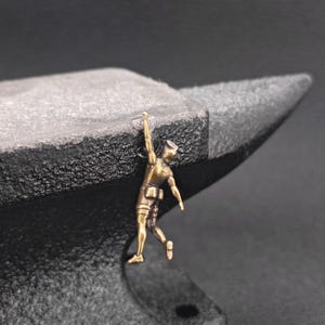 May include: A small bronze-coloured figurine of a climber hanging from the edge of a dark, textured metal anvil. The climber is depicted with one arm extended upwards, gripping the top surface of the anvil, while the body hangs vertically.