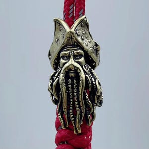 Puede incluir: Una cuenta de metal de color dorado que presenta una representación detallada de un pirata con una barba hecha de tentáculos. La cuenta está unida a un cordón trenzado rojo y blanco.