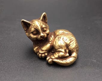May include: A small, golden, brass figurine of a cat curled up sleeping. The cat has a detailed face with large eyes and a small nose.