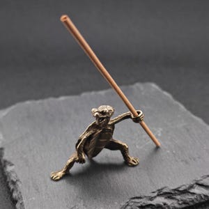 Puede incluir: Un portaincienso de bronce con forma de una pequeña figura agachada que sostiene un palito de incienso marrón. La figura tiene una superficie detallada y texturizada y está colocada sobre una superficie gris oscuro.