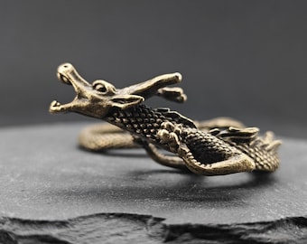 Puede incluir: Un anillo de metal en tono bronce con forma de dragón enroscado con la boca abierta, escamas visibles y detalles texturizados. La cabeza del dragón está situada en la parte delantera y su cuerpo se enrolla para formar la banda del anillo.