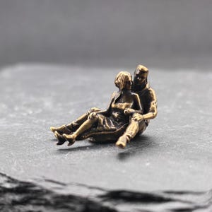 Könnte beinhalten: Eine kleine, antik wirkende Metallfigur in Bronzetönen, die zwei eng beieinander sitzende Personen darstellt. Eine Figur sitzt hinter der anderen und hat die Arme um sie gelegt. Die Skulptur weist eine dunkle, verwitterte Oberfläche auf.