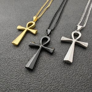 Könnte beinhalten: Drei Ankh-Halsketten in Gold, Schwarz und Silber. Jeder Anhänger hat das traditionelle Design des geschlungenen Kreuzes, das an einer passenden Kette hängt. Die Halsketten werden auf einer strukturierten, dunklen Oberfläche präsentiert.