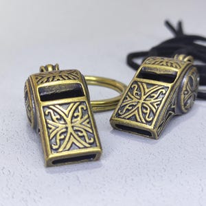 May include: Two antique gold-colored whistles with intricate designs. One whistle is attached to a key ring.