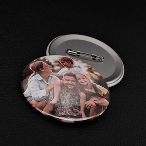 May include: Two round pinback buttons. One button displays a family portrait with a man, woman, and two children. The other button is silver with a safety pin clasp. The buttons are on a black surface.