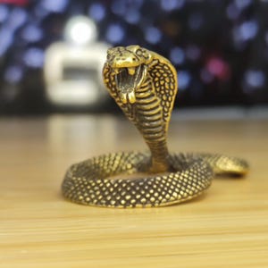 May include: A gold-colored metal figurine of a cobra snake with its hood raised. The snake is coiled with its head facing forward. The figurine is detailed with scales and a textured surface.