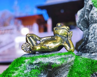 Pode incluir: Uma figurinha de sapo de cor dourada está deitada de costas com os braços cruzados atrás da cabeça. O sapo está descansando sobre uma rocha coberta de musgo verde.