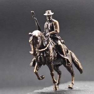Op de afbeelding: Een bronzen beeldje van een man te paard, met een geweer. De ruiter draagt een hoed en zit op een paard met gedetailleerde kenmerken. De sculptuur heeft een vintage esthetiek.