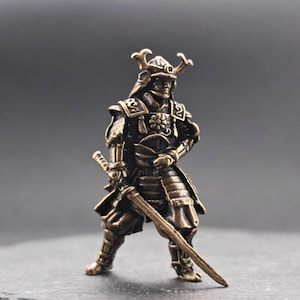 Op de afbeelding: Een bronzen miniatuur samurai krijger beeldje, staand met een zwaard in de hand. De gedetailleerde figuur draagt traditionele harnas en een helm met decoratieve hoorns. Het beeldje heeft een verweerde uitstraling.