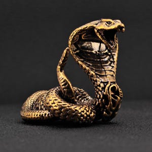 May include: A bronze cobra figurine with an open mouth, ready to strike. The snake's scales and hood are intricately detailed, showcasing a vintage aesthetic. The sculpture is set against a dark background, highlighting its golden hue.