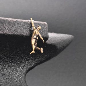 May include: A small, metallic, gold-toned figurine of a person hanging from the edge of a large, textured, black iron anvil. The figure is depicted with one arm extended upward, gripping the top surface of the anvil, while the body hangs down.