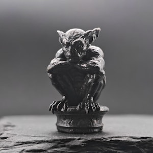 May include: A small, dark grey gargoyle figurine in a seated, hunched position with its arms crossed over its knees. The creature features pointed ears, sharp claws, and a textured, weathered surface, mounted on a circular, pedestal-like base.