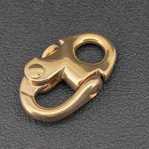 May include: A gold-toned metal lobster clasp featuring a smooth, curved frame with a circular attachment loop at the top. The mechanism includes a small, textured, knurled thumb lever on the side and a flat, circular rivet securing the spring-loaded gate.
