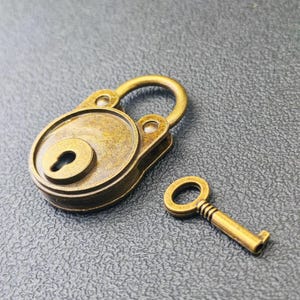 Puede incluir: Un candado de latón de estilo antiguo con una llave a juego. El candado es redondo con un ojo de cerradura y un asa curva. La llave es pequeña y tiene un diseño sencillo. Los artículos están colocados sobre un fondo gris oscuro.