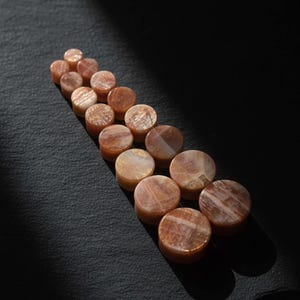 May include: A collection of circular sunstone plugs arranged in a staggered, ascending line. Each piece features a polished, smooth surface with natural orange and brown mineral inclusions that create a shimmering, reflective effect across the stones.