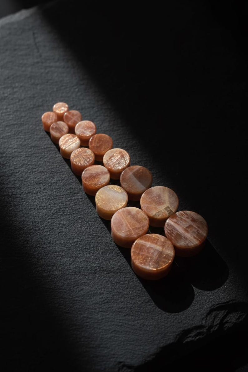 May include: A collection of circular sunstone plugs arranged in a staggered, ascending line. Each piece features a polished, smooth surface with natural orange and brown mineral inclusions that create a shimmering, reflective effect across the stones.