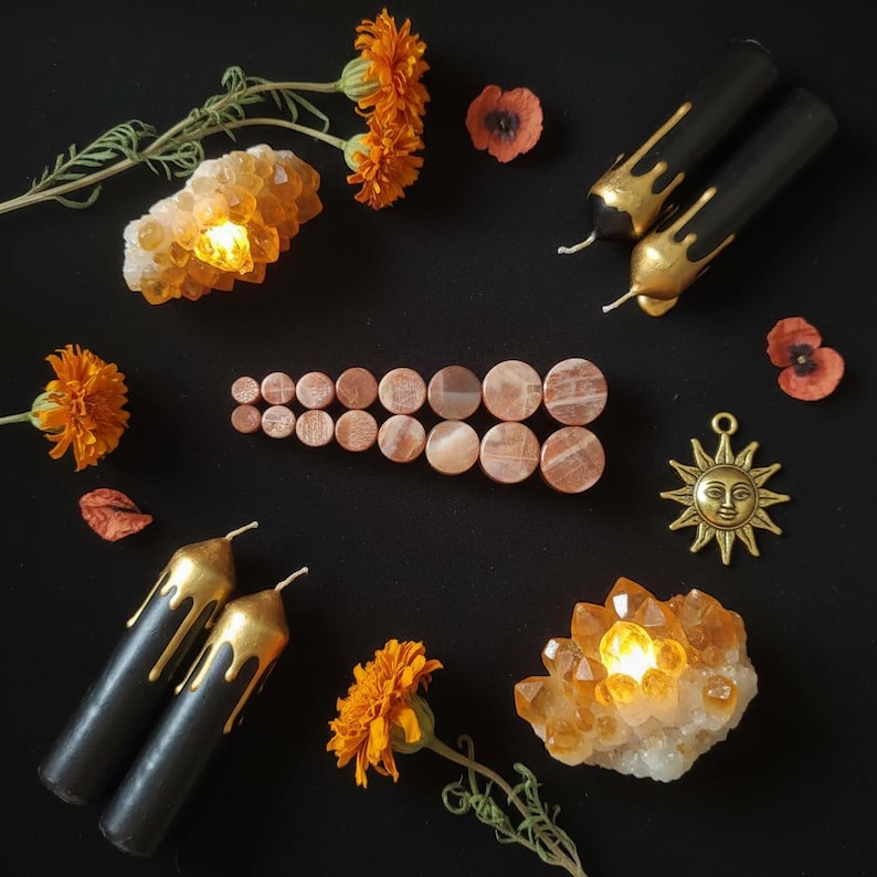 May include: A collection of circular stone ear plugs arranged in a row, surrounded by four black candles with gold drip details, two clusters of glowing orange crystals, orange marigold flowers, red petals, and a gold sun face pendant.