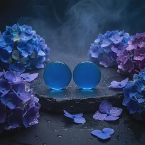 Op de afbeelding: Twee ronde, blauwe glazen objecten liggen op een donker, getextureerd oppervlak, omringd door blauwe en paarse hortensia's. De achtergrond is mistig en donkerblauw, wat een serene en artistieke compositie creëert. De objecten zijn ongeveer 3 cm in diameter.
