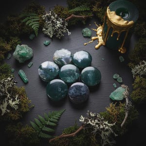 Peut inclure: Une composition sur fond sombre avec des pierres précieuses vertes et bleues, de la mousse et une bougie verte avec des accents dorés. Les pierres sont rondes et présentent des motifs variés. La bougie a de la cire dorée qui coule.