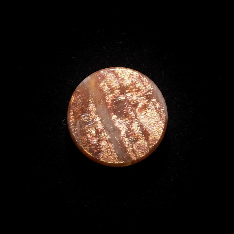 May include: A circular sunstone cabochon featuring a warm, copper-orange base colour with distinct, shimmering metallic inclusions that create a reflective, glittery effect across the surface of the polished, rounded gemstone.