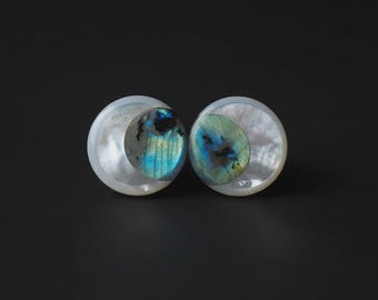 Puede incluir: Dos pendientes redondos con un diseño de media luna de nácar. Cada pendiente presenta una piedra central, más pequeña y redonda, con colores iridiscentes azules y verdes. Los pendientes están sobre un fondo oscuro.