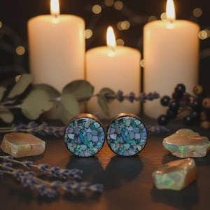 Könnte beinhalten: Zwei runde Ohrstecker mit einem Mosaik aus blauen, grünen und irisierenden Teilen. Die Ohrstecker sind vor dem Hintergrund von brennenden Kerzen, Lavendelzweigen und rohen Edelsteinen platziert, was eine mystische Ästhetik erzeugt.