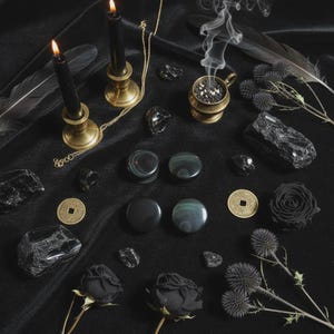 Puede incluir: Una composición de objetos místicos sobre una tela negra. Incluye velas negras encendidas en candelabros de latón, piedras negras, un quemador de incienso humeante, rosas negras y monedas decorativas. También hay una cadena dorada y plumas negras.