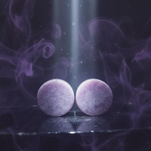 Puede incluir: Dos objetos redondos de color lavanda con una superficie texturizada son el centro de la imagen. El fondo presenta humo púrpura arremolinado y un haz de luz. Los objetos parecen ser pendientes o piedras decorativas.
