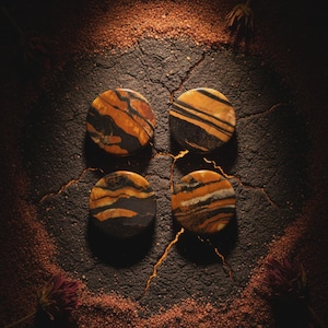 Op de afbeelding: Vier ronde, gepolijste stenen met een opvallend patroon van zwarte en oranje-bruine strepen en wervelingen. De stenen zijn gerangschikt op een donkere, getextureerde ondergrond met een verspreiding van fijne, roodbruine korrels.