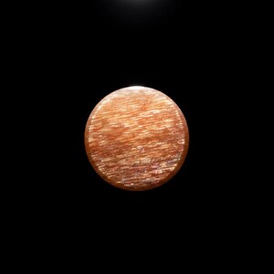 May include: A circular sunstone cabochon featuring a warm orange and copper colour palette. The surface displays a distinct chatoyant effect with fine, parallel, shimmering inclusions that create a reflective, fibrous texture across the stone.