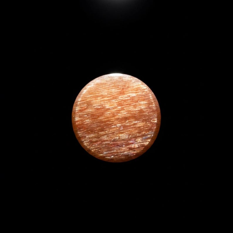 May include: A circular sunstone cabochon featuring a warm orange and copper colour palette. The surface displays a distinct chatoyant effect with fine, parallel, shimmering inclusions that create a reflective, fibrous texture across the stone.