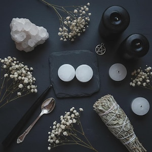 Op de afbeelding: Een bovenaanzicht van een verzameling spirituele items op een donkere ondergrond. De opstelling omvat zwarte kaarsen, witte kwartskristallen, een zilveren lepel, een saliebundel en kleine witte kaarsen. Delicate witte bloemen zijn door de compositie verspreid.