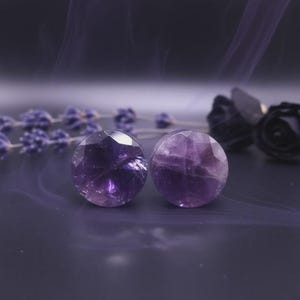 Puede incluir: Dos tapones redondos de amatista facetada. Los tapones son de color púrpura intenso y tienen una superficie pulida. El fondo es de color púrpura oscuro con ramitas de lavanda y rosas oscuras.