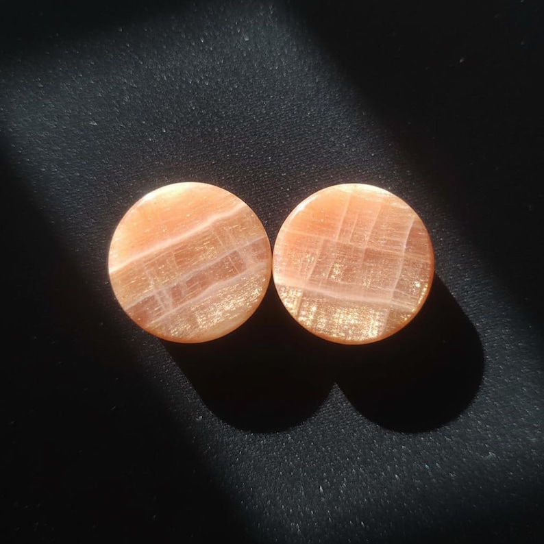 May include: Two circular ear plugs made of polished peach-coloured stone with a translucent quality. The surface of each plug displays a natural grid-like pattern of lighter, intersecting lines and a subtle shimmering effect across the smooth texture.