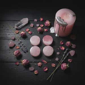 Puede incluir: Una vela rosa con goteo plateado, rodeada de cuatro tapones de piedra rosa, rosas secas, cristales y ramitas de lavanda sobre una superficie de madera oscura. La disposición evoca temas espirituales.