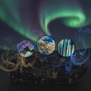 May include: Three polished labradorite stones, each with a unique pattern of iridescent colours including blue, purple, and gold. The stones rest on a dark, textured surface, with a backdrop of a starry night and aurora borealis.