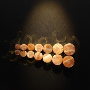May include: A collection of fourteen circular sunstone ear plugs arranged in two rows of seven, increasing in size from left to right. Each plug features a polished, translucent orange-brown surface with natural mineral inclusions and shimmer.