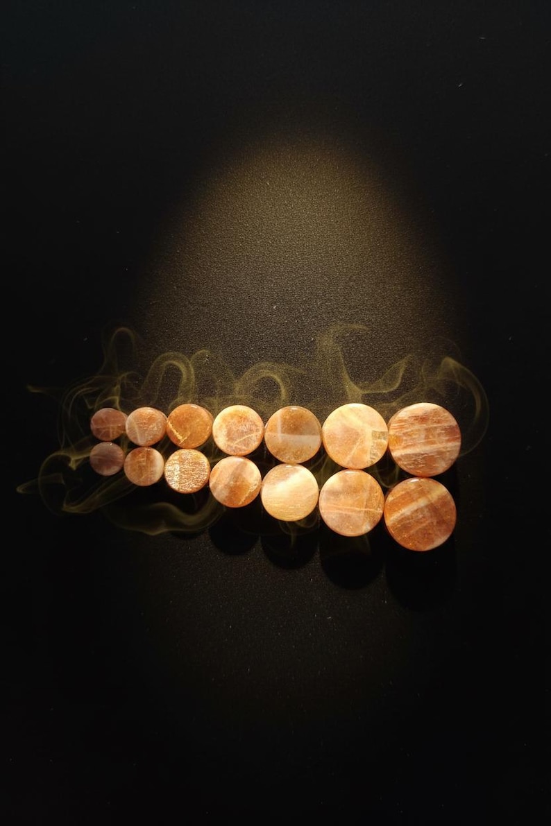 May include: A collection of fourteen circular sunstone ear plugs arranged in two rows of seven, increasing in size from left to right. Each plug features a polished, translucent orange-brown surface with natural mineral inclusions and shimmer.