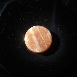 May include: A circular sunstone cabochon featuring a warm orange and peach hue with internal shimmering inclusions. The polished gemstone displays a reflective, glittery surface texture with subtle white linear veining across the centre.