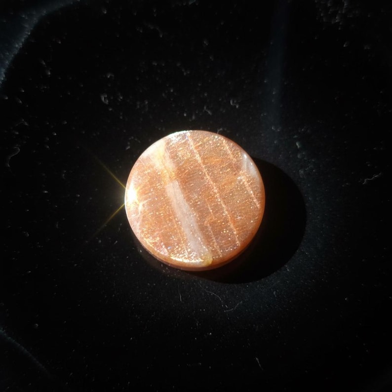 May include: A circular sunstone cabochon featuring a warm orange and peach hue with internal shimmering inclusions. The polished gemstone displays a reflective, glittery surface texture with subtle white linear veining across the centre.