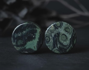 Puede incluir: Dos tapones redondos de piedra verde oscuro con patrones únicos. Los tapones tienen una superficie pulida y una mezcla de remolinos y manchas verdes oscuros y negros. El fondo es oscuro, enfatizando los colores y texturas.
