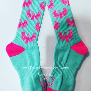 May include: A pair of teal blue socks with pink butterfly designs. The socks have a pink heel and toe. The socks have the text "Little Angels" on the bottom of the socks. The text "World Down Syndrome Day Apparel" and "Order today $12" is printed on the socks.