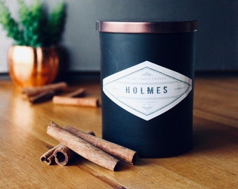 May include: A black candle jar with a copper lid and a white label that reads "Pure Soy Candles Holmes Hand Poured in NY". The candle is sitting on a wooden surface with cinnamon sticks.
